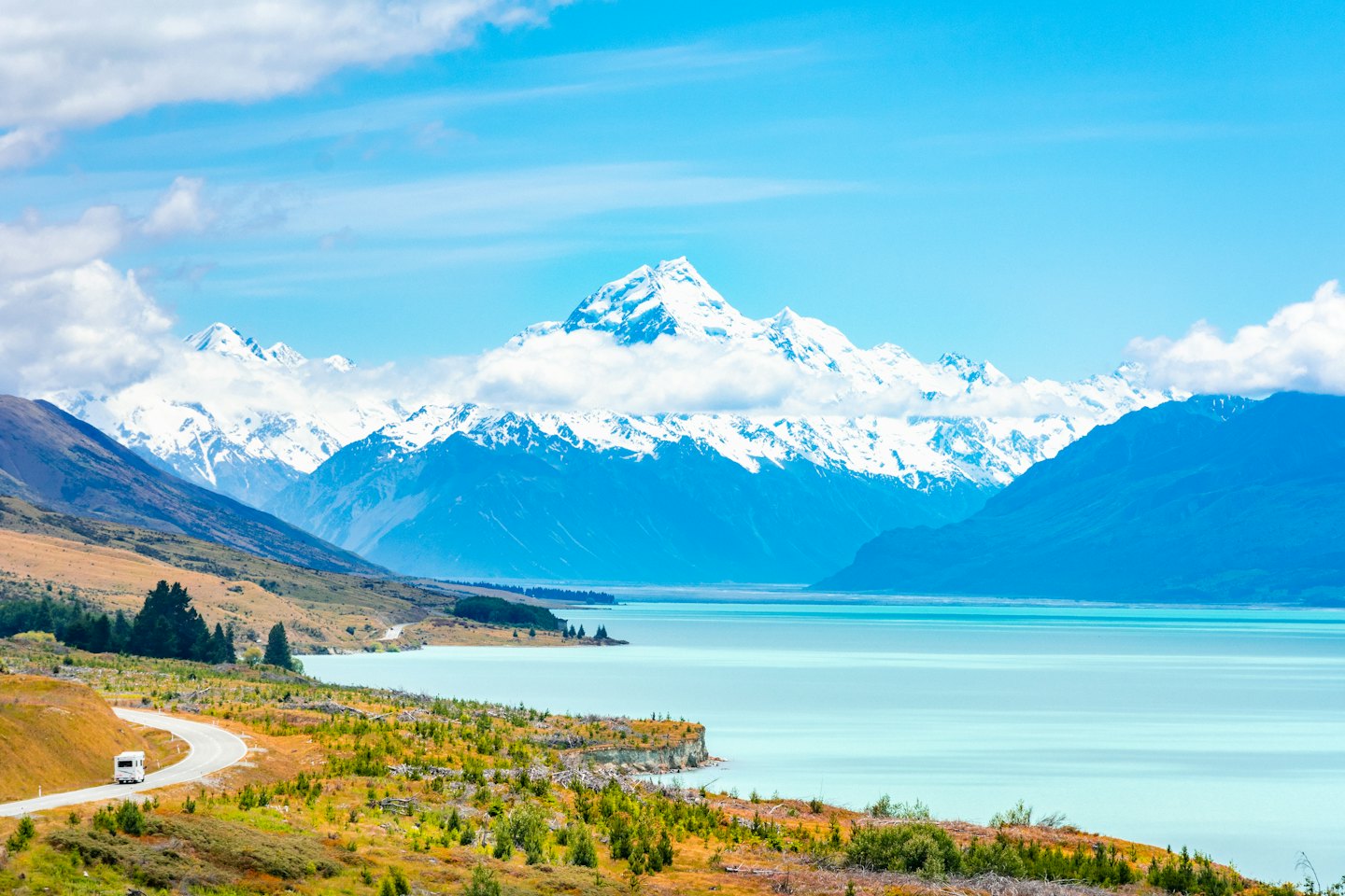 A white recreational vehicle drives along a lake near snow-capped mountains.
