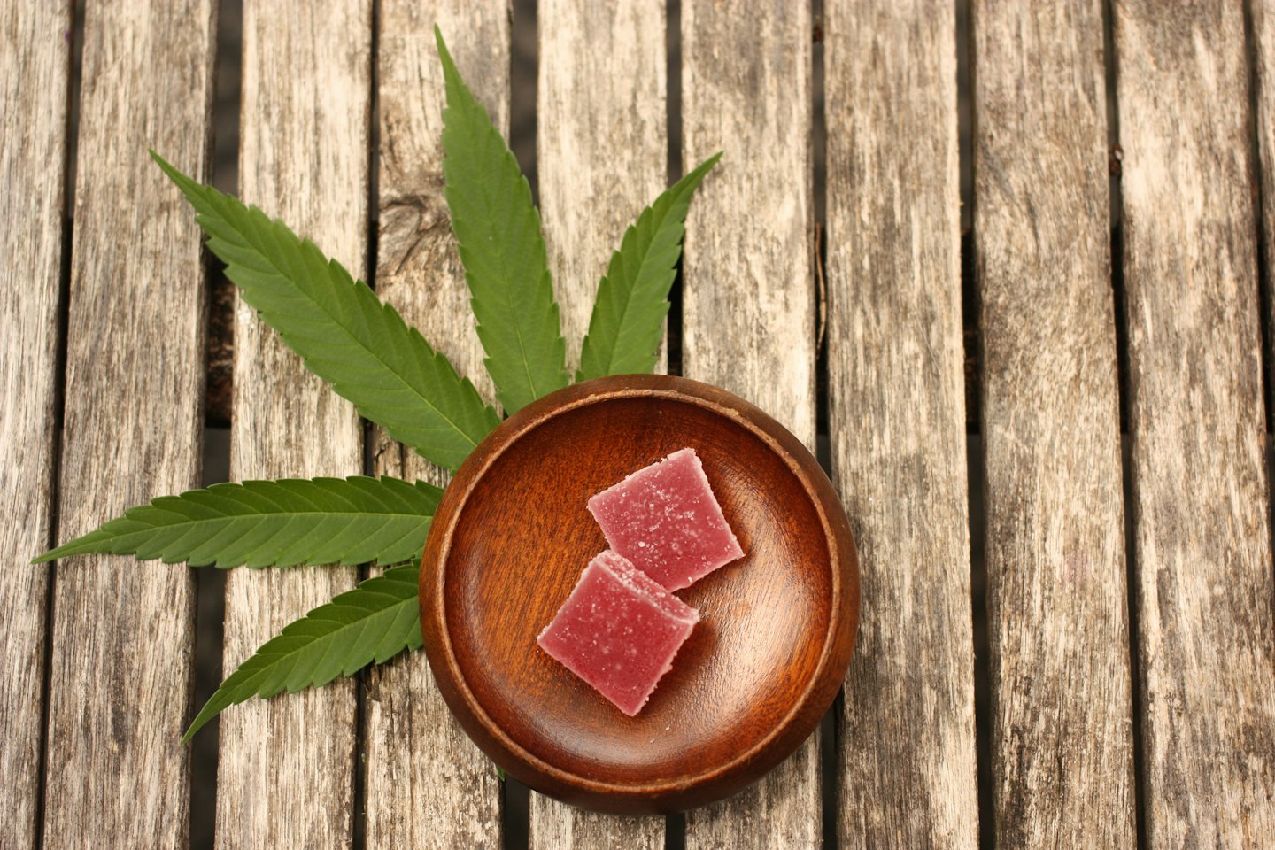 Two sugar-coated pink gummies sit in a wooden bowl with cannabis leaves.